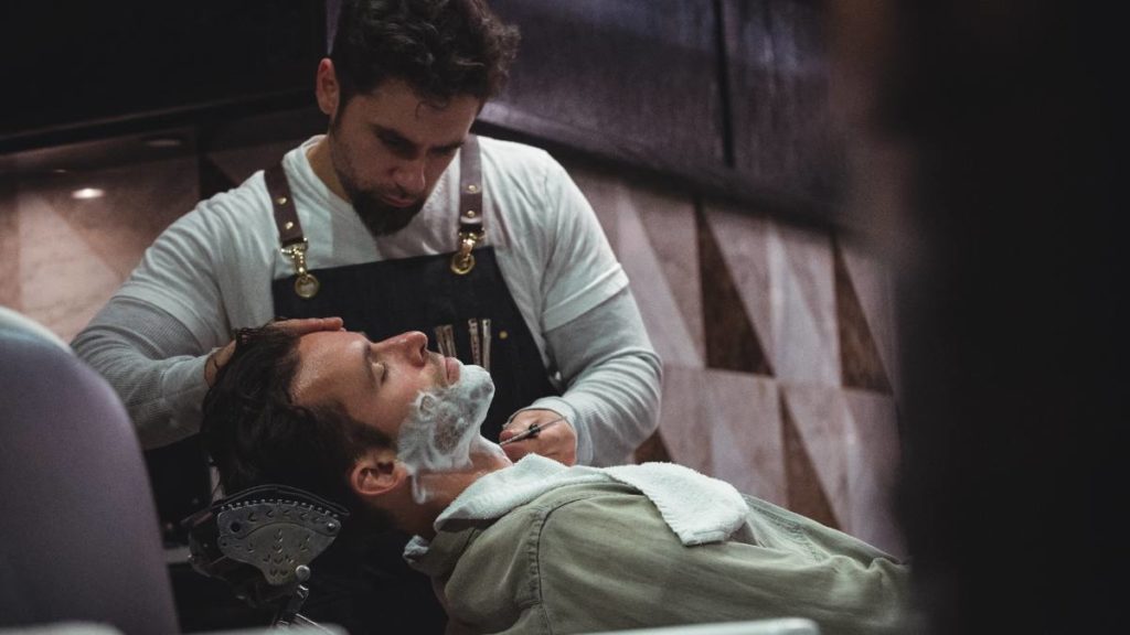 Barbeiro a barbear a barba de um cliente com espuma numa barbearia
