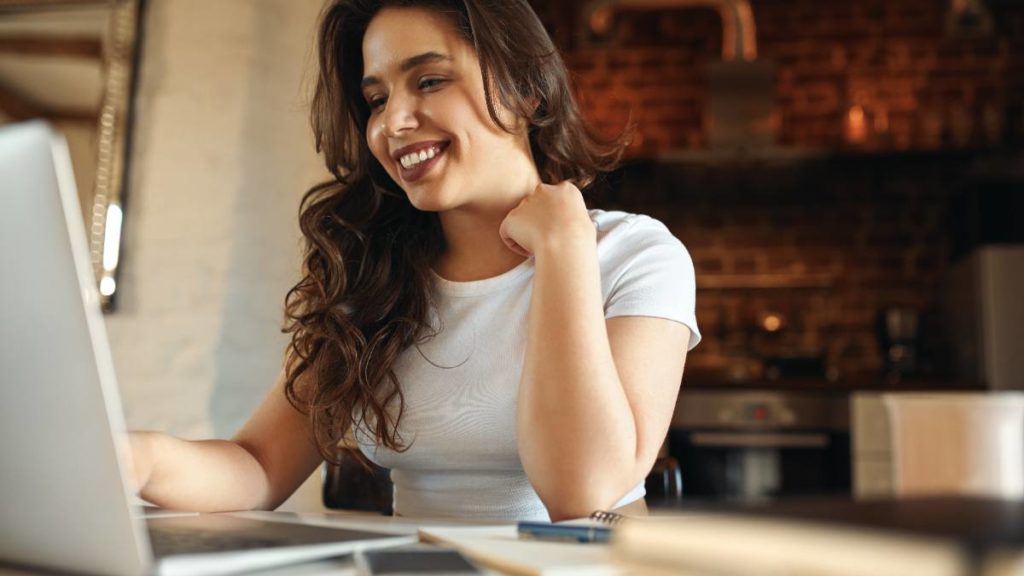 Mulher sorridente a estudar num computador portátil numa cozinha, com um caderno de apontamentos ao lado
