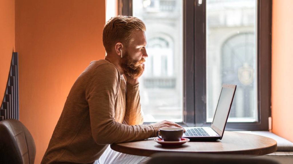 Homem sentado a uma mesa junto a uma janela, a trabalhar num computador portátil com uma chávena de café ao lado