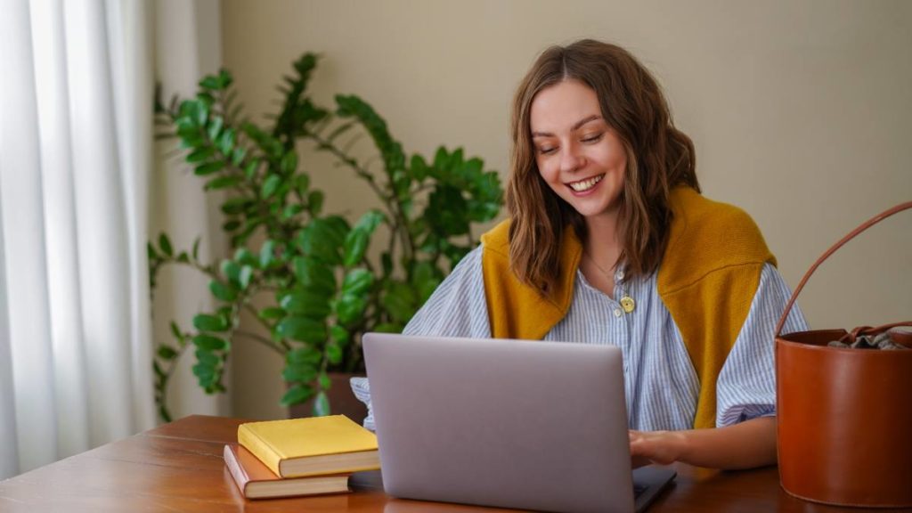 Mulher sorridente a estudar num computador portátil em casa, com livros e uma planta ao lado
