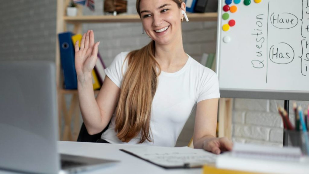 Mulher a acenar durante uma aula de inglês online, com um quadro branco com vocabulário atrás