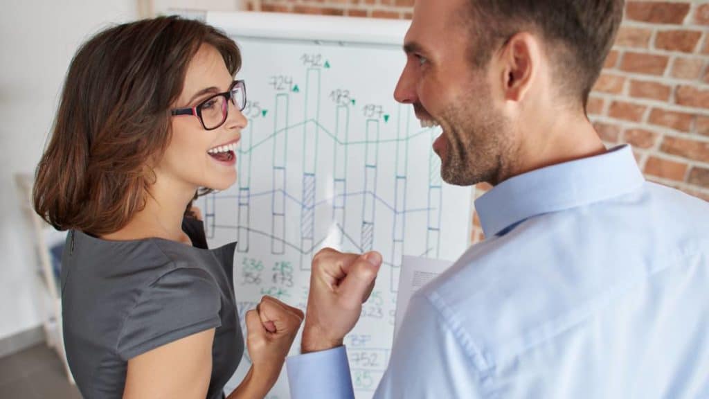 Homem e mulher a celebrar com os punhos fechados junto a um quadro branco com gráficos de vendas