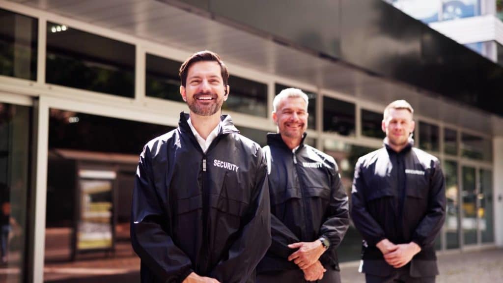 Três seguranças/porteiros de uniforme a sorrir junto à entrada de um edifício