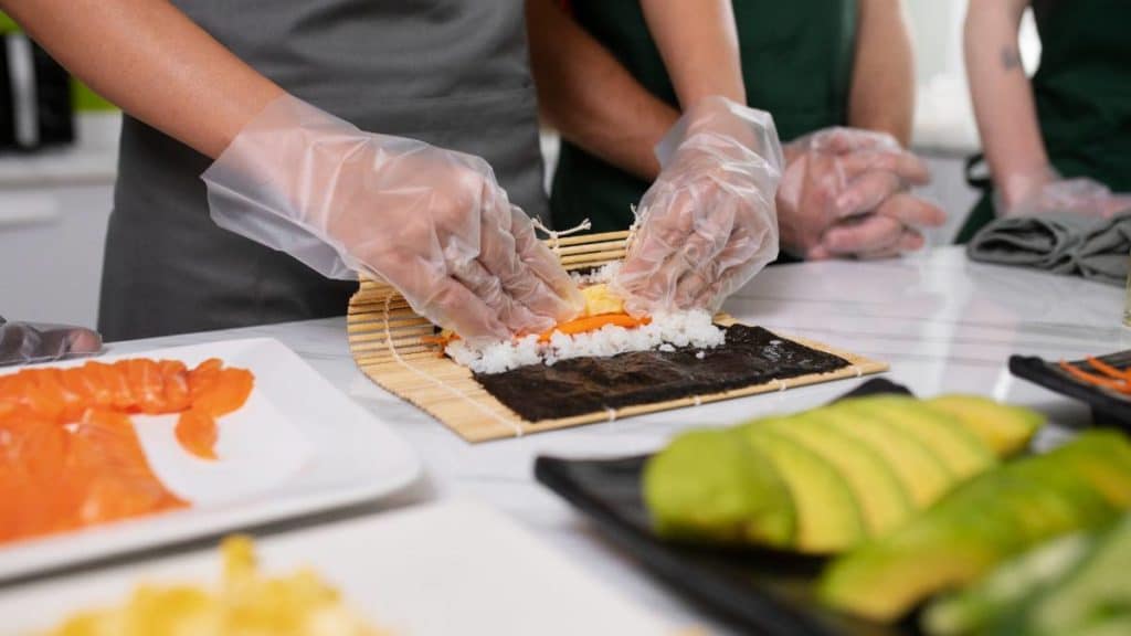 Mãos com luvas a preparar um rolo de sushi sobre uma esteira de bambu, com salmão e legumes ao lado