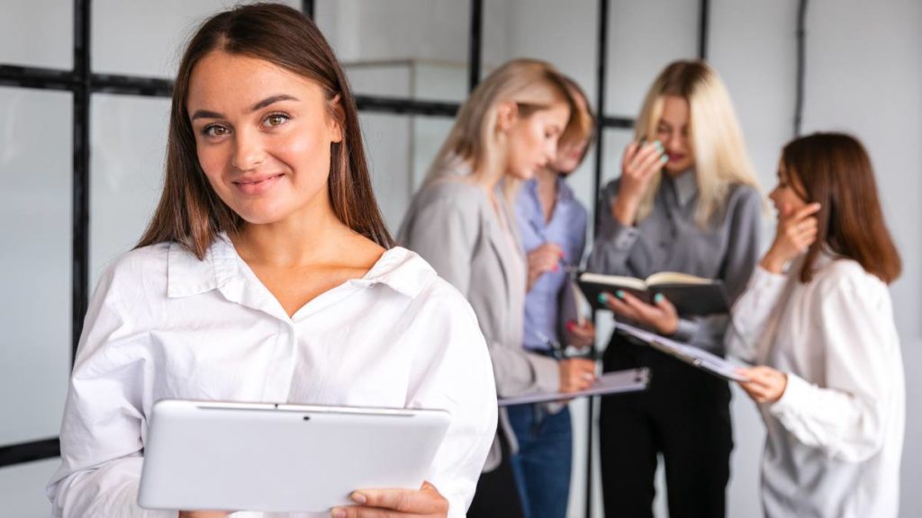 Técnica administrativa com um tablet, com colegas a trabalhar em equipa num escritório ao fundo