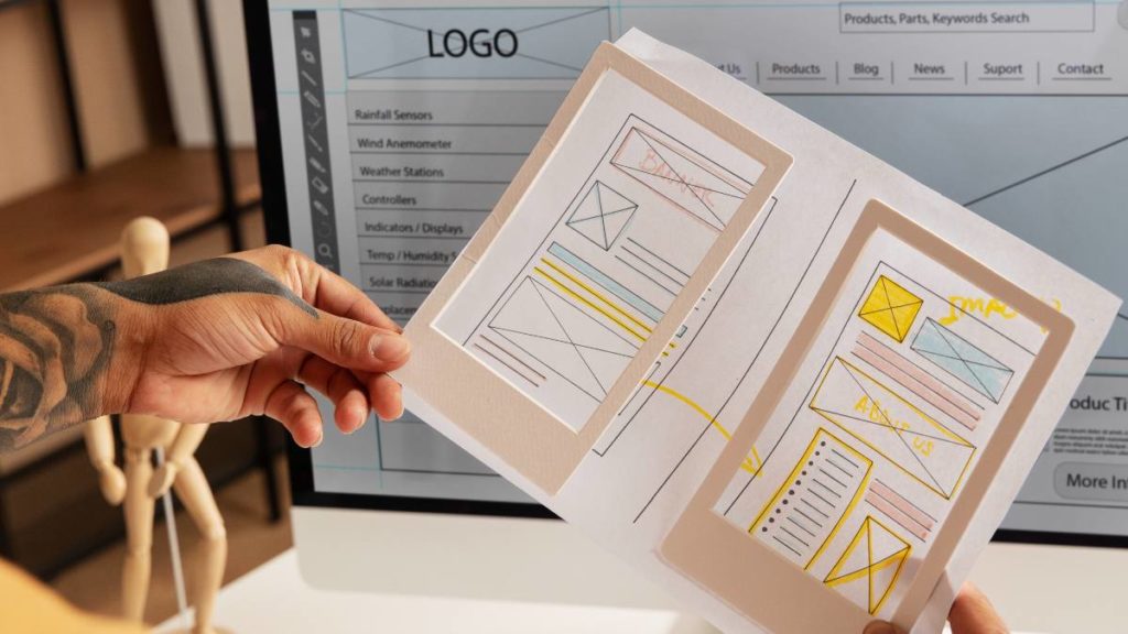 Mãos a segurar esboços em papel de wireframes de um website, com o design digital ao fundo num ecrã